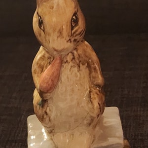 May include: A ceramic figurine of Peter Rabbit, a brown rabbit with long ears, holding a carrot in its paws. The rabbit is sitting on a small, white platform with a brown base.