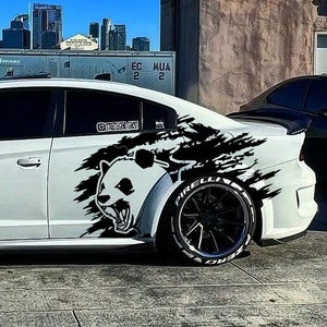 May include: White Dodge Charger with a black panda graphic on the side. The car has black rims with white lettering on the tires. The background includes a cityscape and a clear blue sky.