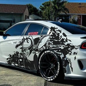 May include: A white Dodge Charger with black accents and custom graphics. The car features black rims with white lettering, a rear spoiler, and a black and white graphic design on the side. The car is parked on a paved driveway.