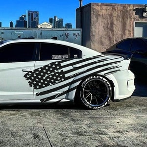 May include: A white Dodge Charger with a black and white American flag decal on the side. The car is parked on a rooftop with a city skyline in the background.