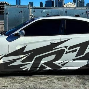 May include: White Dodge Charger with black and gray SRT graphics on the side. The car has black rims with white lettering on the tires. The car is parked on a paved surface with other vehicles in the background.