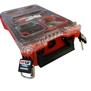 Può includere: Una cassetta degli attrezzi Milwaukee Packout rossa con un coperchio trasparente e una maniglia nera. La cassetta ha un'etichetta che dice "M12 ProPex Expander".