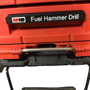 May include: A red tool case with a black label displaying "M18 Fuel Hammer Drill" in white text. The case features a metal handle and a black base. The M18 logo is in red and white.