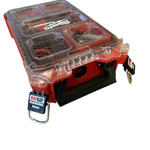 Op de afbeelding: Rode Milwaukee Packout gereedschapskist met een doorzichtig deksel en meerdere compartimenten. De kist heeft een zwarte handgreep en een metalen ring met een zwart label met de tekst "M12 Installation Driver".