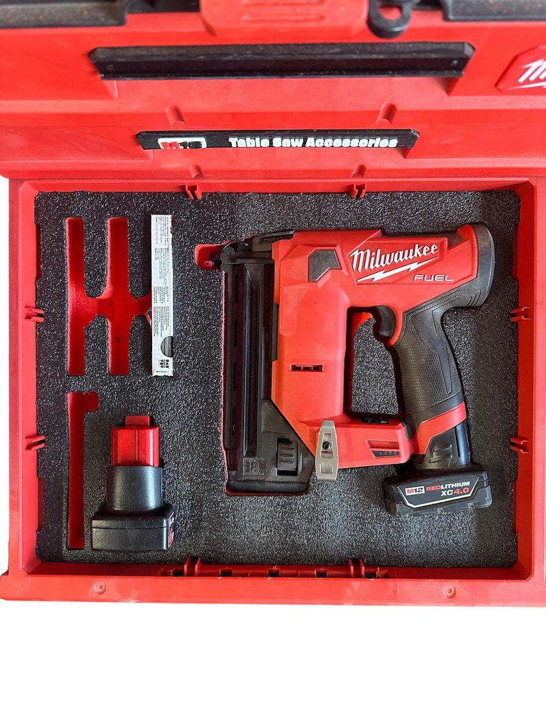 K&ouml;nnte beinhalten: Ein roter Milwaukee FUEL 18-Gauge-Brad-Nagler mit einem schwarzen Akku und einem schwarzen Transportkoffer. Der Koffer tr&auml;gt den Schriftzug "Table Saw Accessories" auf der Oberseite.