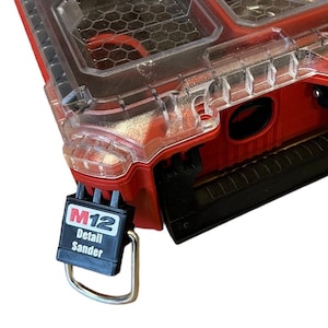 Può includere: Una cassetta degli attrezzi Milwaukee rossa con un coperchio in plastica trasparente. La cassetta ha una maniglia nera e un'etichetta nera e argentata M12 Detail Sander attaccata ad essa.