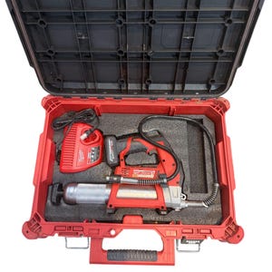 May include: A red and black power tool kit with a black lid. The kit contains a red grease gun, a red battery charger, and a black battery. The case is open, revealing the tools nestled in black foam.