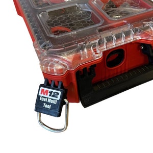 Könnte beinhalten: Eine rote Milwaukee Packout Werkzeugbox mit einem durchsichtigen Deckel und einem schwarzen M12 Fuel Multi Tool Etikett, das an dem Griff befestigt ist.
