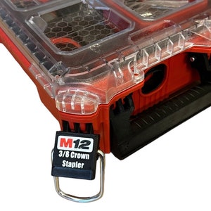 Può includere: Una cassetta degli attrezzi Milwaukee Packout rossa con un coperchio trasparente. La cassetta ha un cinturino nero con un anello di metallo e un'etichetta nera che dice "M12 3/8 Crown Stapler".