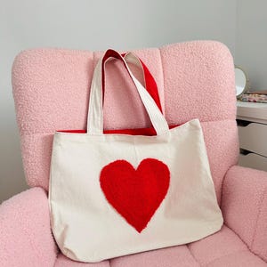 May include: A white canvas tote bag with a red fuzzy heart on the front. The bag has red handles and is sitting on a pink fuzzy chair.