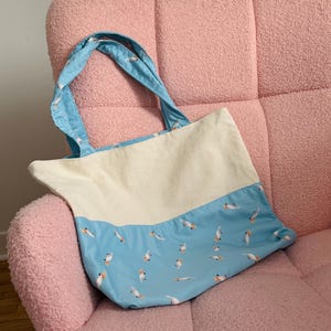 May include: A white and light blue tote bag with a pattern of small white birds on the light blue portion. The bag has two blue handles.