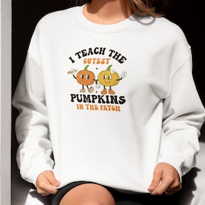 Puede incluir: Sudadera blanca con un gráfico de dos calabazas de dibujos animados tomándose de la mano. El texto dice "Enseño las calabazas más lindas del huerto."