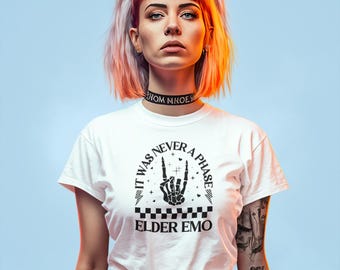 Może przedstawiać: Kobieta z różowymi i białymi włosami nosi biały t-shirt z czarnym nadrukiem, na którym widnieje napis "It Was Never A Phase" i "Elder Emo". Nadruk przedstawia szkieletową dłoń wykonującą rockowy gest.