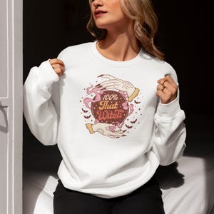 May include: White sweatshirt with a graphic of a hand holding a crystal ball with the text "100% That Witch" in a retro font.