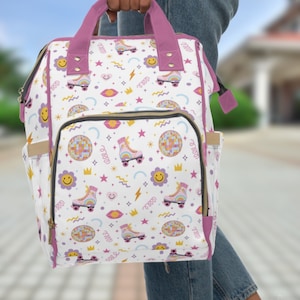 Könnte beinhalten: Eine weiße Wickeltasche mit einem rosafarbenen Griff und rosafarbenen Akzenten. Die Tasche hat ein Muster aus Rollschuhen, Regenbögen, Smiley-Gesichtern, Sternen und Discokugeln.