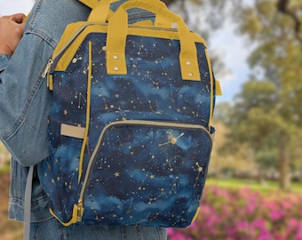 Puede incluir: Una mochila azul y dorada con estampado de constelaciones. La mochila tiene correas amarillas y un bolsillo con cremallera en la parte delantera.