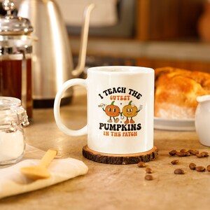 Könnte beinhalten: Weiße Keramik-Kaffeetasse mit einem braunen Holz-Untersetzer. Die Tasse hat ein Motiv mit zwei lächelnden Kürbissen mit dem Text "Ich unterrichte die süßesten Kürbisse im Feld".