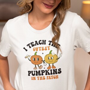 Puede incluir: Camiseta blanca con el texto "I TEACH THE CUTEST PUMPKINS IN THE PATCH". La camiseta presenta un diseño retro de dos calabazas de dibujos animados, una con un lápiz y la otra haciendo el signo de la paz. Las calabazas son naranjas y amarillas.