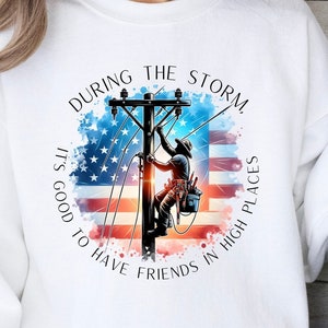 May include: White sweatshirt with a graphic of an electrician working on a power line with an American flag in the background. The text reads "During the storm, it's good to have friends in high places."