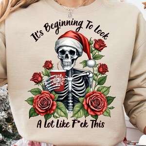 May include: A beige sweatshirt with a skeleton wearing a Santa hat holding a mug. The text on the sweatshirt reads "It's Beginning To Look A Lot Like F*ck This".