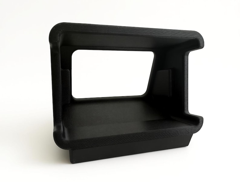 Pu&ograve; includere: Un oggetto nero rettangolare stampato in 3D con una superficie strutturata. Presenta un ampio spazio rettangolare aperto al centro, con una base a forma di mensola. L'oggetto ha angoli arrotondati e un design angolare robusto.