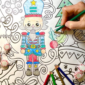 Pode incluir: Uma página de colorir com uma figura central de quebra-nozes colorida em azul, vermelho e amarelo, rodeada por motivos natalícios como uma árvore de Natal, folhas de azevinho e espirais. Vários lápis de cor e rebuçados estão presentes.