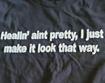May include: Black t-shirt with white text that reads "Healin' aint pretty, I just make it look that way."