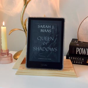 May include: A Kindle e-reader displaying the book "Queen of Shadows" by Sarah J. Maas, on a wooden stand. A lit candle in a pink holder and a decorative lamp with a pink glass shade are also visible.