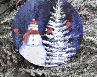 May include: A round, flat ornament featuring a painted winter scene with a snowman wearing a red hat, scarf, and mittens standing next to a white, snow-covered evergreen tree. A small red bird sits on the tree, all set against a dark blue background.