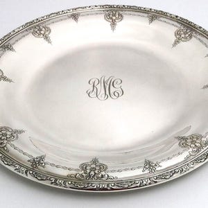Pode incluir: Travessa oval de servir em prata com uma borda ornamentada com motivos florais e volutas. O centro da travessa apresenta iniciais gravadas. A borda tem um padrão detalhado e repetitivo. Uma peça clássica para servir comida.