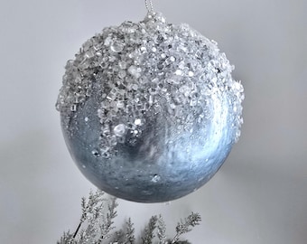 Puede incluir: Una bola de Navidad plateada con una parte superior texturizada similar a un cristal y un acabado metálico liso. La bola cuelga de un hilo plateado, y debajo se ve una ramita de vegetación helada. Una decoración festiva para la temporada navideña.