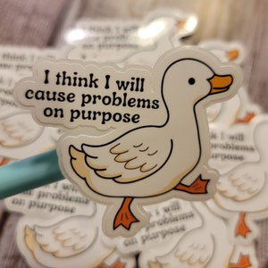 May include: A white duck sticker with orange feet and beak, and the text "I think I will cause problems on purpose" in a speech bubble. The sticker is on a wooden surface with other similar stickers.