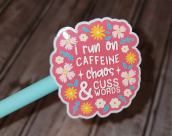 May include: A pink sticker with a white border and floral accents. The sticker reads "I run on caffeine + chaos + & cuss words" in white text. Yellow, white, and blue flowers decorate the sticker.