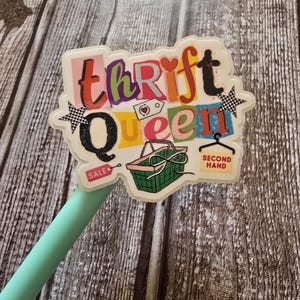 May include: A colorful sticker with the words "Thrift Queen" in a playful font. The design includes a shopping basket, stars, and the words "Sale" and "Second Hand". The sticker is on a light green pencil.