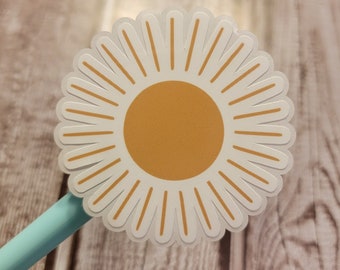 May include: A white and orange daisy sticker with a scalloped edge. The sticker has a bright orange center and white petals with orange lines. A light blue pen is partially visible in the image.