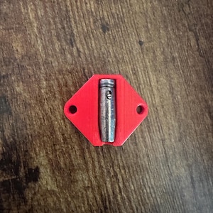 May include: A red, diamond-shaped object with two holes and a silver metal cylinder in the center. The cylinder has a small, engraved "e" on its side. The object is likely a component or part of a larger mechanism.