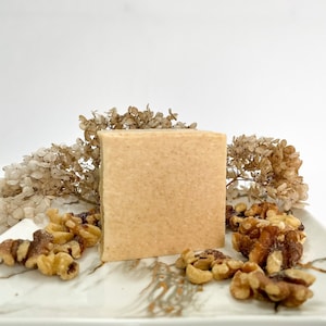 May include: A light brown bar of soap with a textured surface, sitting on a white plate with walnuts scattered around it. The soap is surrounded by dried flowers.