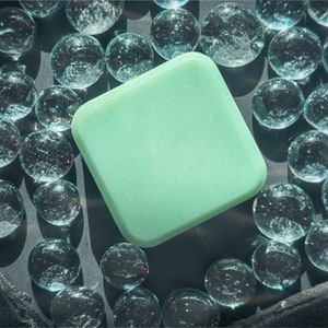 May include: A square, light green object with rounded corners is surrounded by clear glass marbles. The marbles reflect light, creating a sparkling effect. The objects are arranged on a dark surface.