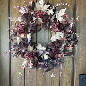 Peut inclure: Une couronne circulaire composée d'un mélange de feuillage bordeaux foncé et beige atténué, incluant des feuilles en forme d'érable et des grappes de petites baies rouge profond. L'arrangement est dense, avec des textures variées.