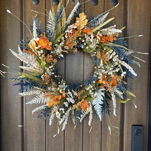 Peut inclure: Une couronne circulaire composée d'un mélange de feuillage orange, bleu et blanc. L'arrangement comprend des feuilles orange, des brins bleu foncé semblables à des fougères, des panaches blancs plumeux et de fines herbes séchées, le tout entrelacé.