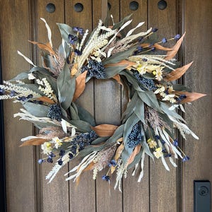May include: A circular wreath composed of dried foliage, featuring long sage green leaves, tan-coloured dried leaves, clusters of small dark blue berries, and wispy cream-coloured grasses. The arrangement is accented with small dark blue and white flowers.
