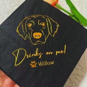 Pode incluir: Guardanapo de papel preto com um contorno dourado da cabeça de um cão e o texto "Drinks on me! Willow"