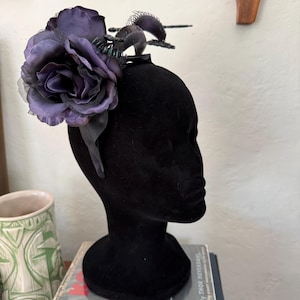 Può includere: Un cerchietto nero caratterizzato da una grande rosa in tessuto viola scuro su un lato e una piccola figura di creatura nera, spinosa e simile a un drago, posizionata sulla parte superiore. Il cerchietto è esposto su un manichino nero.