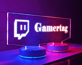 May include: A purple and red illuminated sign with the Twitch logo and the word "Gamertag" on a white background. The sign is on a white base with a blue light.