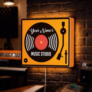 Op de afbeelding: Een vierkant, verlicht bord met een draaitafelontwerp. Het bord heeft een oranje achtergrond met een zwart-witte plaatgrafiek en de tekst "Your Name's MUSIC STUDIO". Het is gemonteerd op een bakstenen muur.
