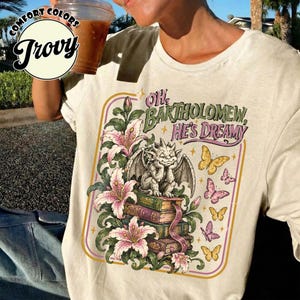 Op de afbeelding: Een crèmekleurig T-shirt met een grafische print van een waterspuwer op een stapel boeken, omringd door lelies en vlinders. De tekst "OH, BARTHOLOMEW, HE'S DREAMY" staat in een gestileerd lettertype. Het ontwerp heeft een dunne rand.