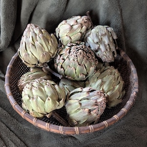 May include: A woven basket filled with fresh artichokes. The artichokes are a muted green color with textured leaves. The basket is made of a light brown woven material. The background is a dark green, textured fabric.