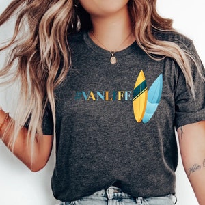 Peut inclure: Une femme portant un t-shirt gris fonc&eacute; avec le texte "#VANLIFE" et un graphique de deux planches de surf. Elle porte un jean bleu clair et des chaussures blanches &agrave; enfiler.