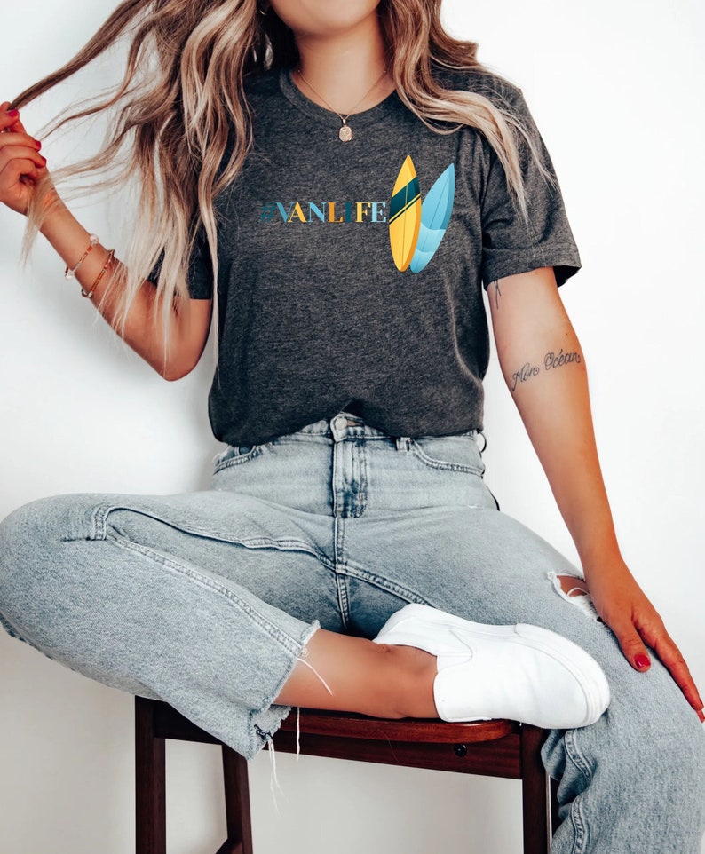 Peut inclure: Une femme portant un t-shirt gris fonc&eacute; avec le texte "#VANLIFE" et un graphique de deux planches de surf. Elle porte un jean bleu clair et des chaussures blanches &agrave; enfiler.