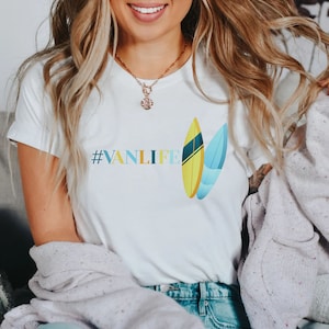 Peut inclure: T-shirt blanc avec le texte "#VANLIFE" et un graphique de deux planches de surf. Les planches de surf sont jaunes, bleues et vertes.